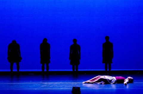 A dancer lies on the floor lit in a pool of blue and white down light. Four other dancers stand upstage in silhouette from a blue cyclorama.