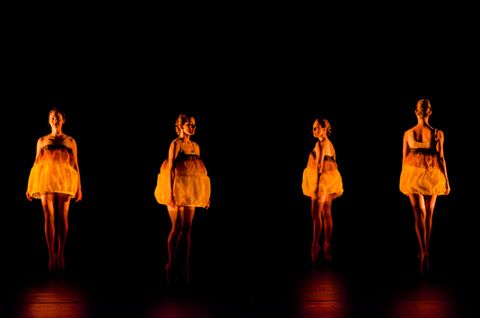 Four dancers en pointe with gold dresses. They are lit in amber light from mid thy up. The dancers represent lanterns floating down a river.