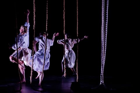 Three dancers appear in a black void among a forest of dangling ropes. They are wearing all white and are lit by cool lavender side light.