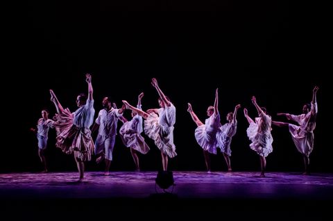 Multiple dancers appear as though they are in a blck void. They wear white blouses and ruffled skirts. They are lit by a cool high breakup and lavender back light.