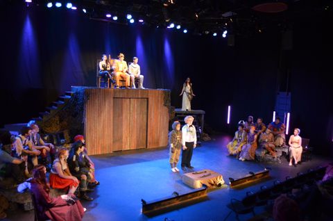 A wide view of the stage for Dionysus of the Holocaust during a perfomance. The races are divide in two and sit on opposite sides of the stage. The Gods look down on them as they state their cases. The Gods are lit in bright, warm light.