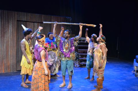 Several people of a tribe standing around a person at center. The center person is holding a staff above his head. The poeple standing around him hold a closed fist in the air as symbol of power.