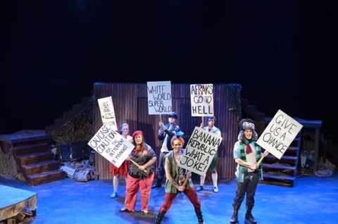 The Caucases stand with signs in protest of the Afracks in Dionysus of the Holocaust. They are lit in a blue down light with a cool white breakup pattern.