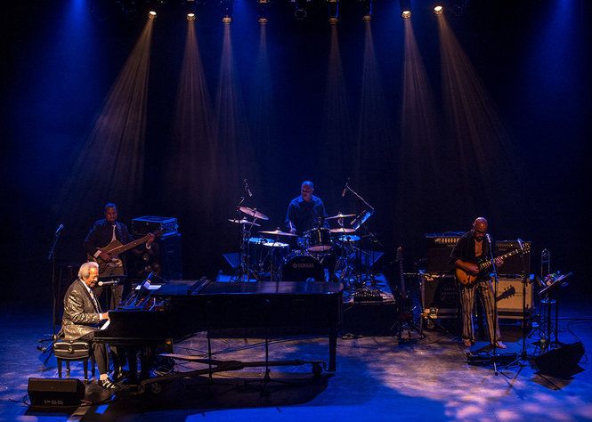 Wide view of the stage for Allen Toussaint concert. The stage is lit with blue back light. Allen Toussaint is lit with a front special and the other musicians are lit with a warm breakup from upstage. Shafts of light an be seen through the haze.