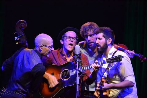 The band members of Steel Wheels group around a center vocal microphone. They are lit by blue and warm white back light and a front warm special.