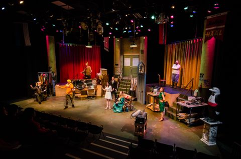 Wide view of the stage for Dove during a performance. It is the back stage of a theatre with an entrance up center and a stage on both sides of the space. We see dressing room mirrors and storage.