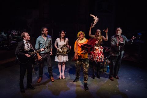 Sevral actors/musicians stand in a line with a look of shock as a dead dove falls to the ground.